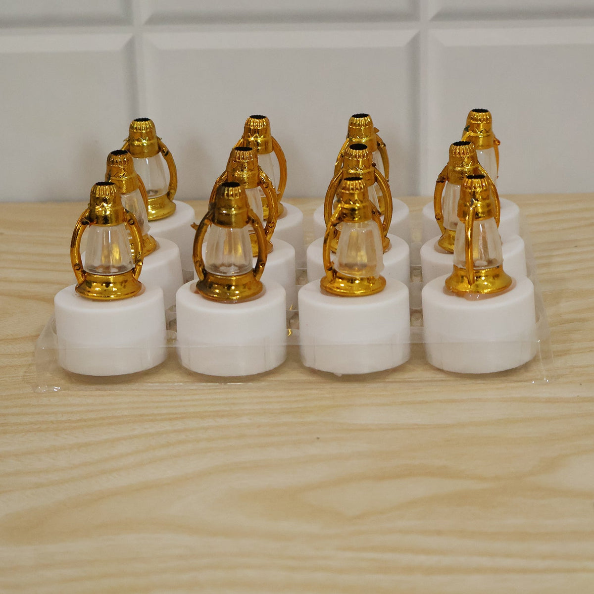 Twelve golden lantern LED candles with white bases are neatly packaged in a clear plastic tray on a light wooden surface.
