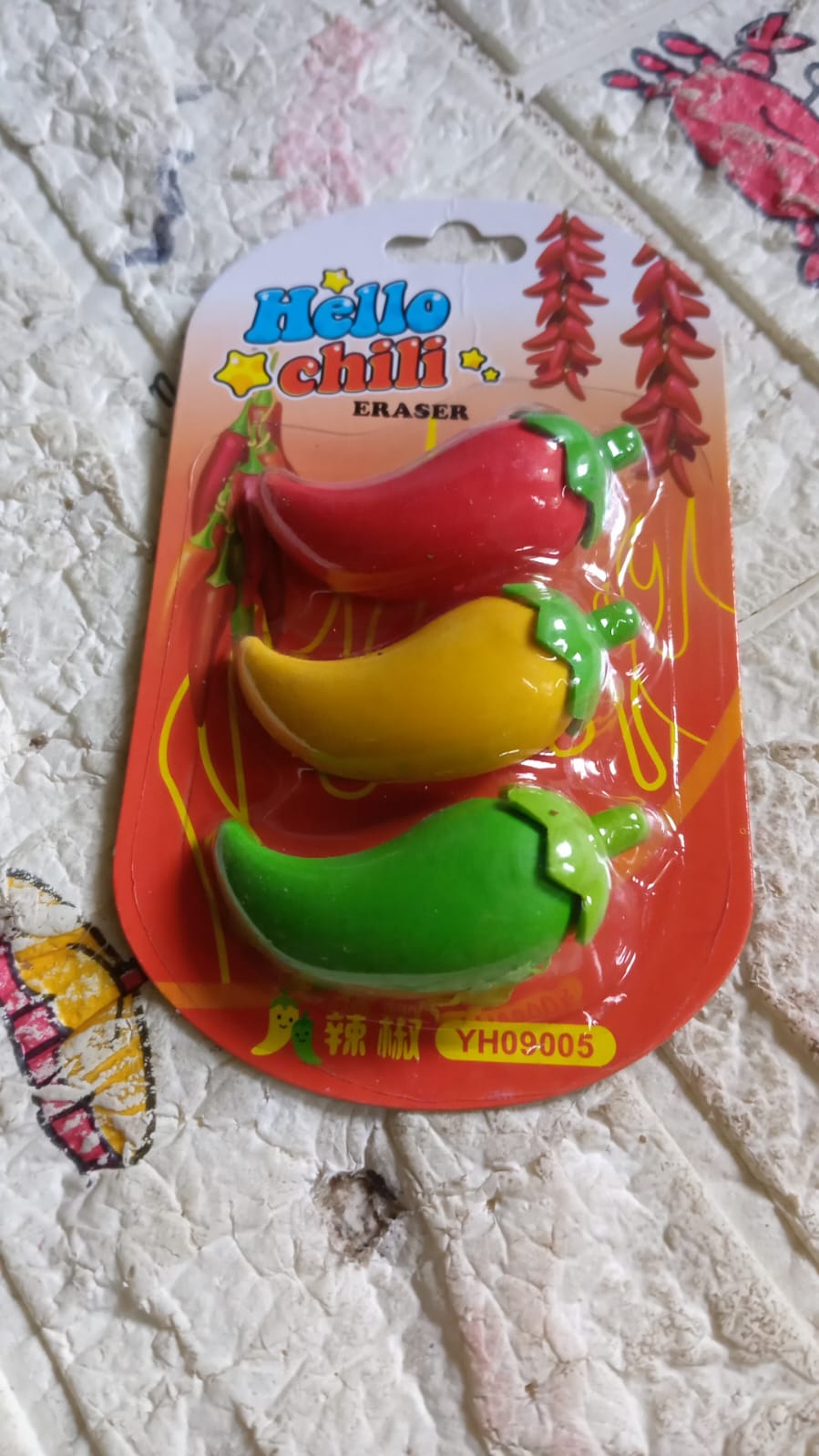 A blister pack of three chili pepper erasers in red, yellow, and green, resting on a textured, light-colored surface.