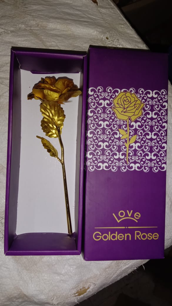 An open purple gift box containing a gold-plated rose, next to its lid, on a textured dark surface.