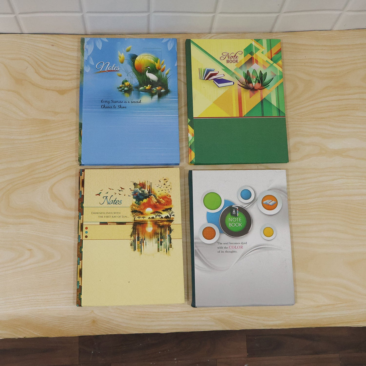 Four different hardbound ruled notebooks with colorful covers featuring various designs, arranged on a light wooden surface.
