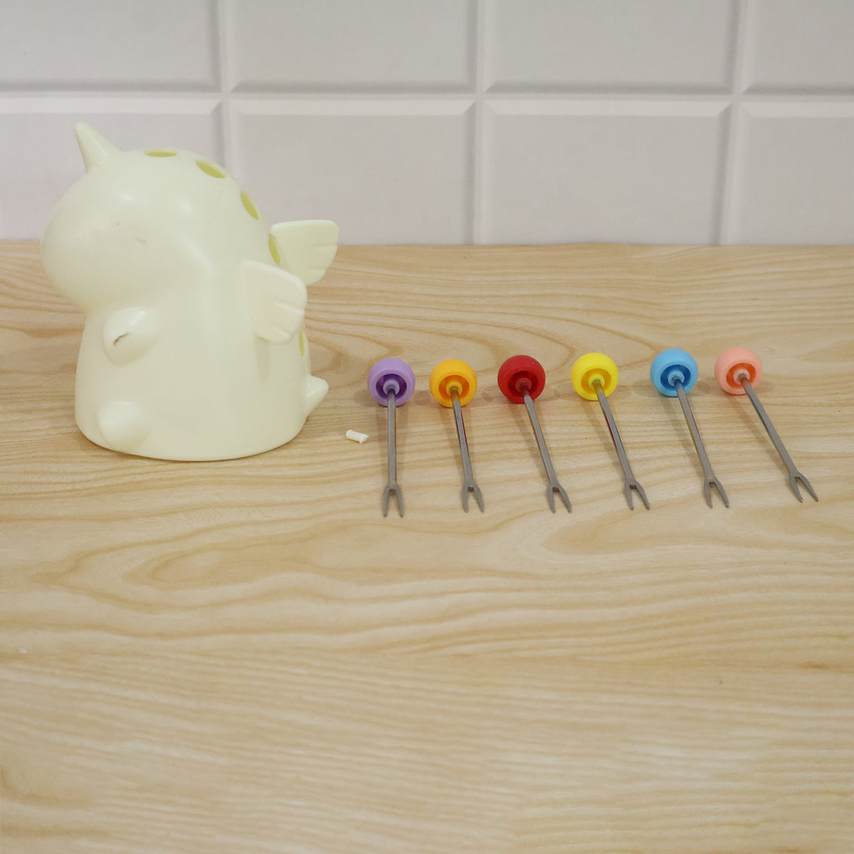 A cream unicorn stand sits beside six colorful fruit forks laid out on a light wooden table against a white tiled wall.