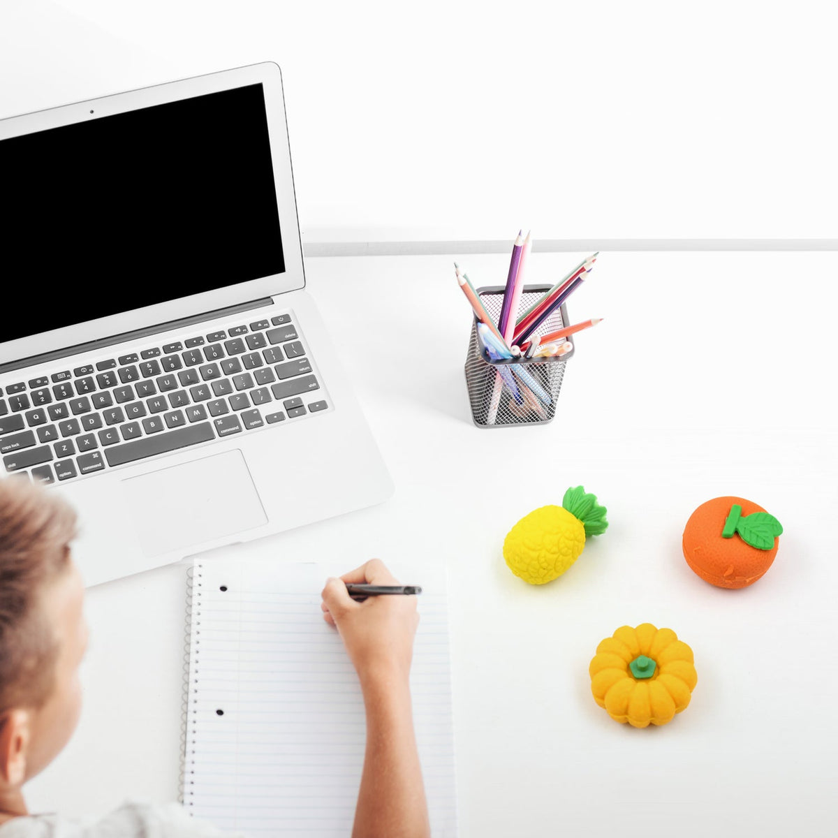 A person writes in a notebook next to a laptop and a pencil holder, with fruit-shaped erasers on the white desk.