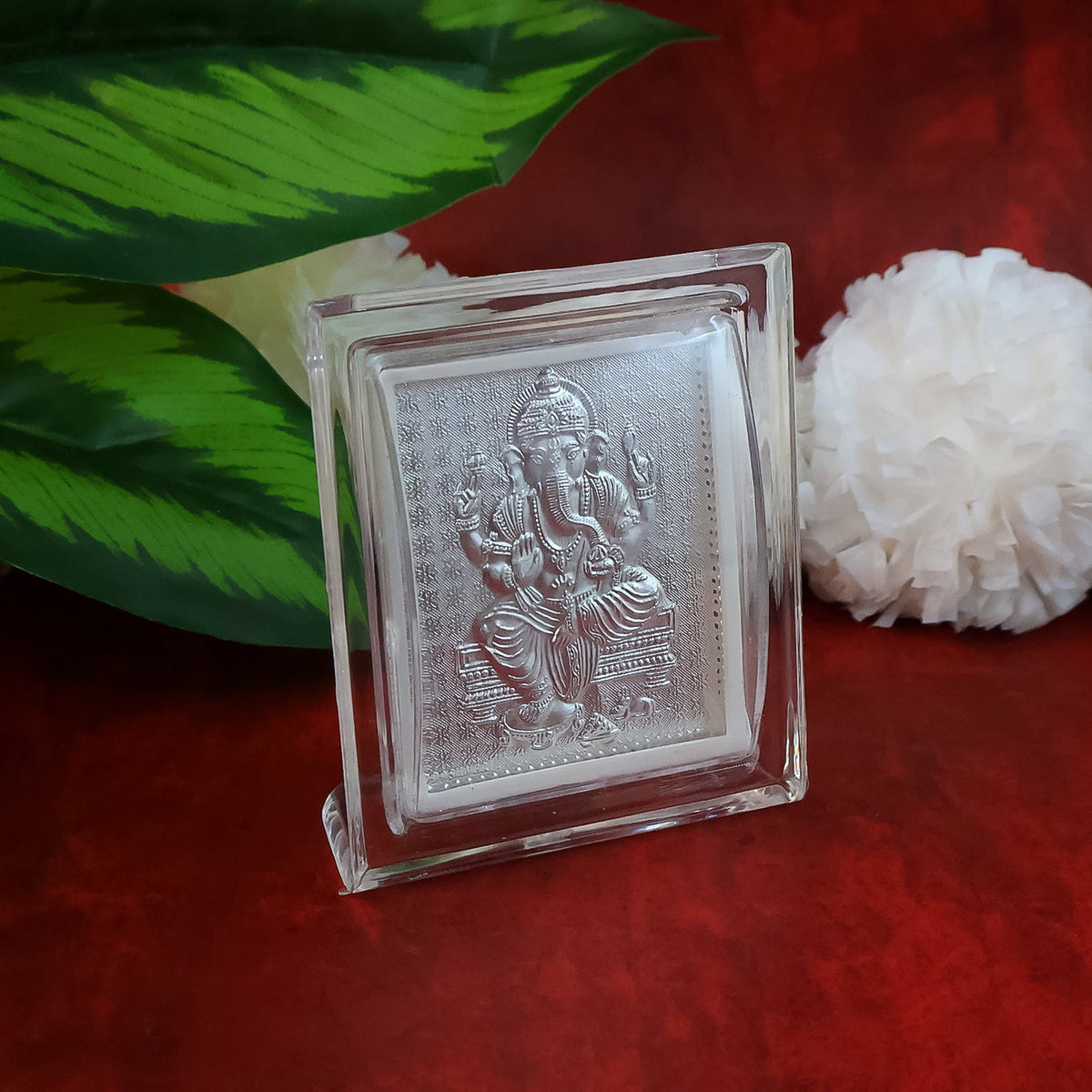 Silver-plated Ganesha idol in an acrylic frame, with a white fluffy decoration and green leaves on a red surface.