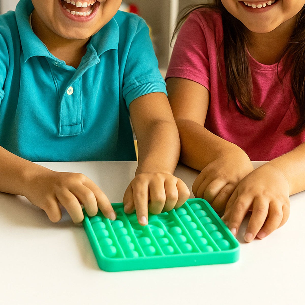 Two children's hands pressing bubbles on a square green silicone pop-it fidget toy on a white table.