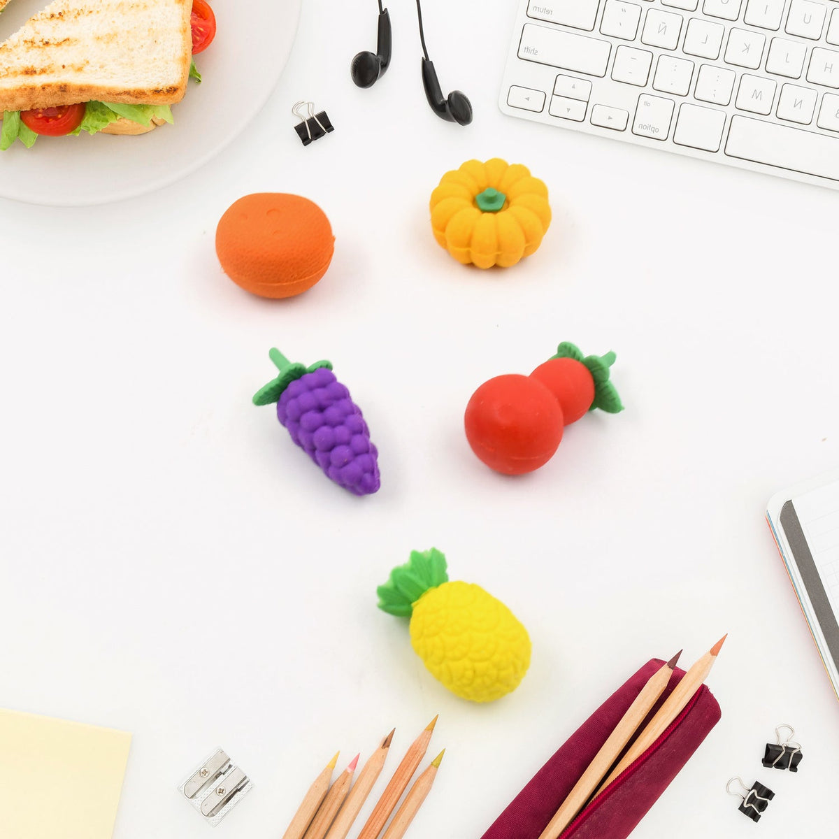 Assorted fruit-shaped erasers, including an orange, pumpkin, grapes, cherry, and pineapple, are scattered on a white desk.
