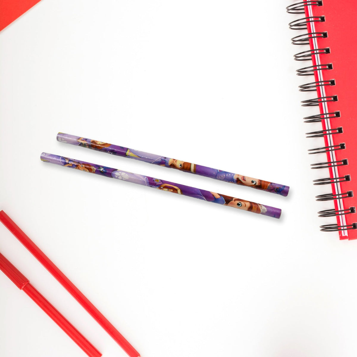 Two purple Sofia the First pencils lying on a white surface next to a red spiral notebook and red pens.
