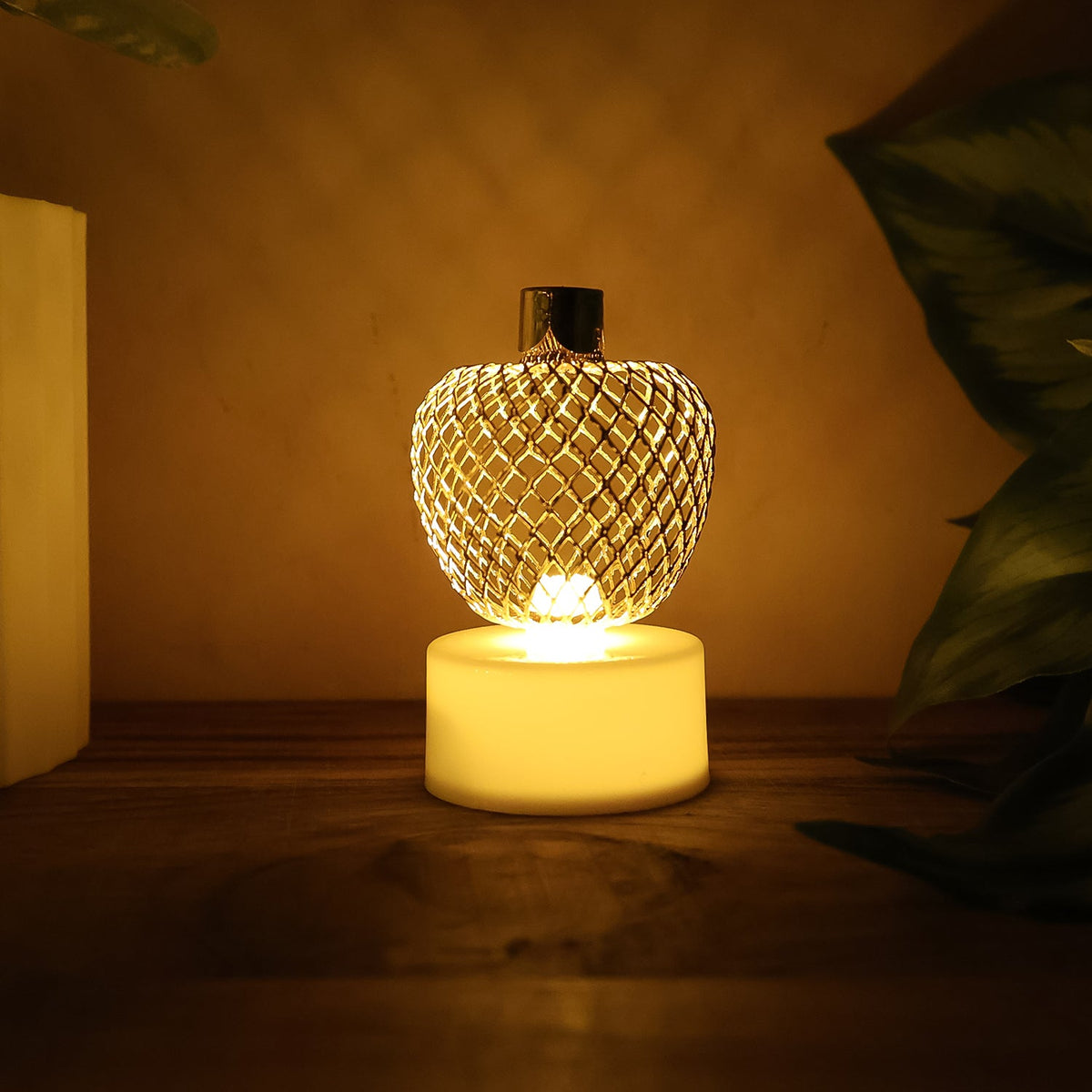 A single glowing gold mesh LED tealight on a wooden surface, with a green plant leaf partially visible on the right.