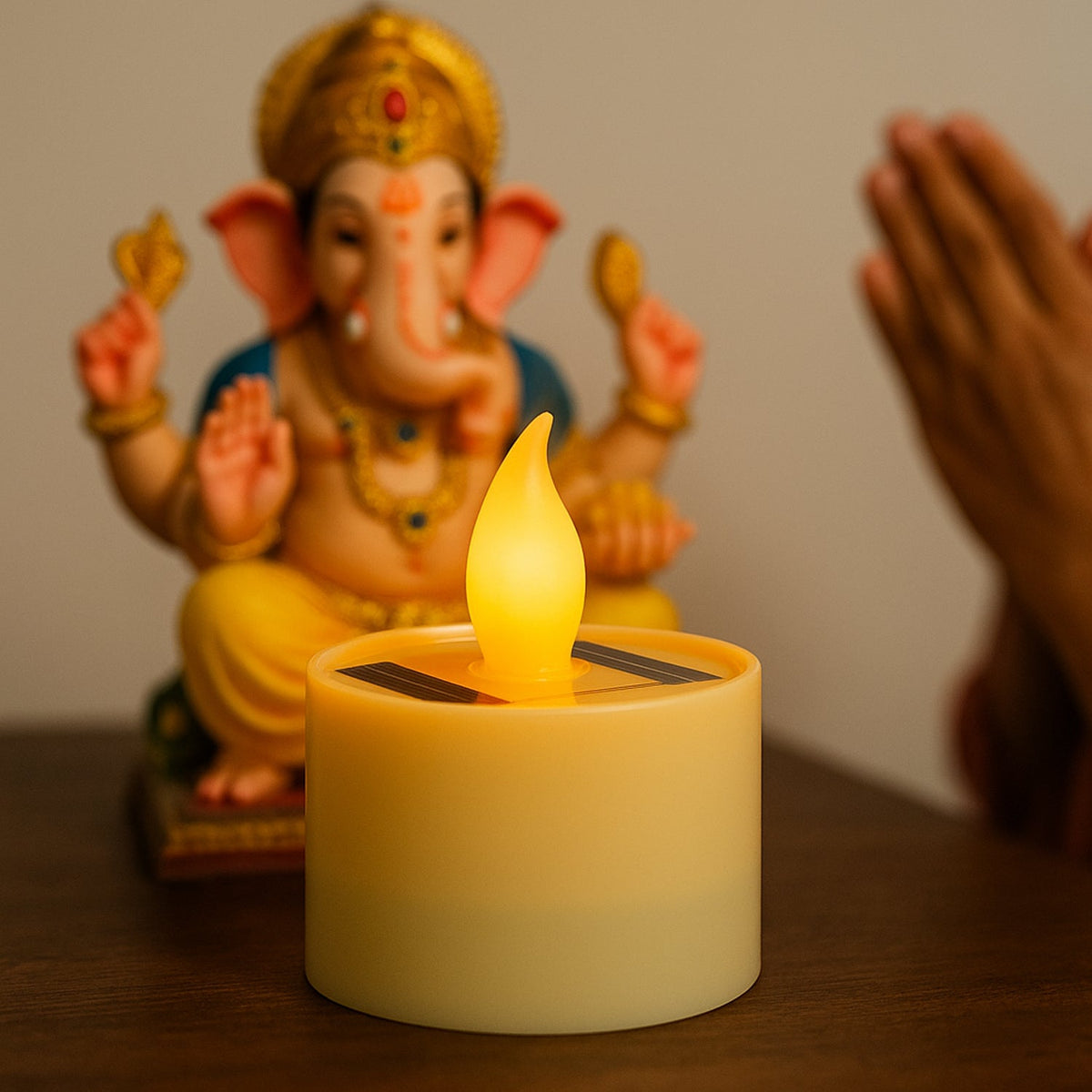 A glowing solar LED candle lamp on a wooden surface, with a blurred Ganesha statue and praying hands in the background.