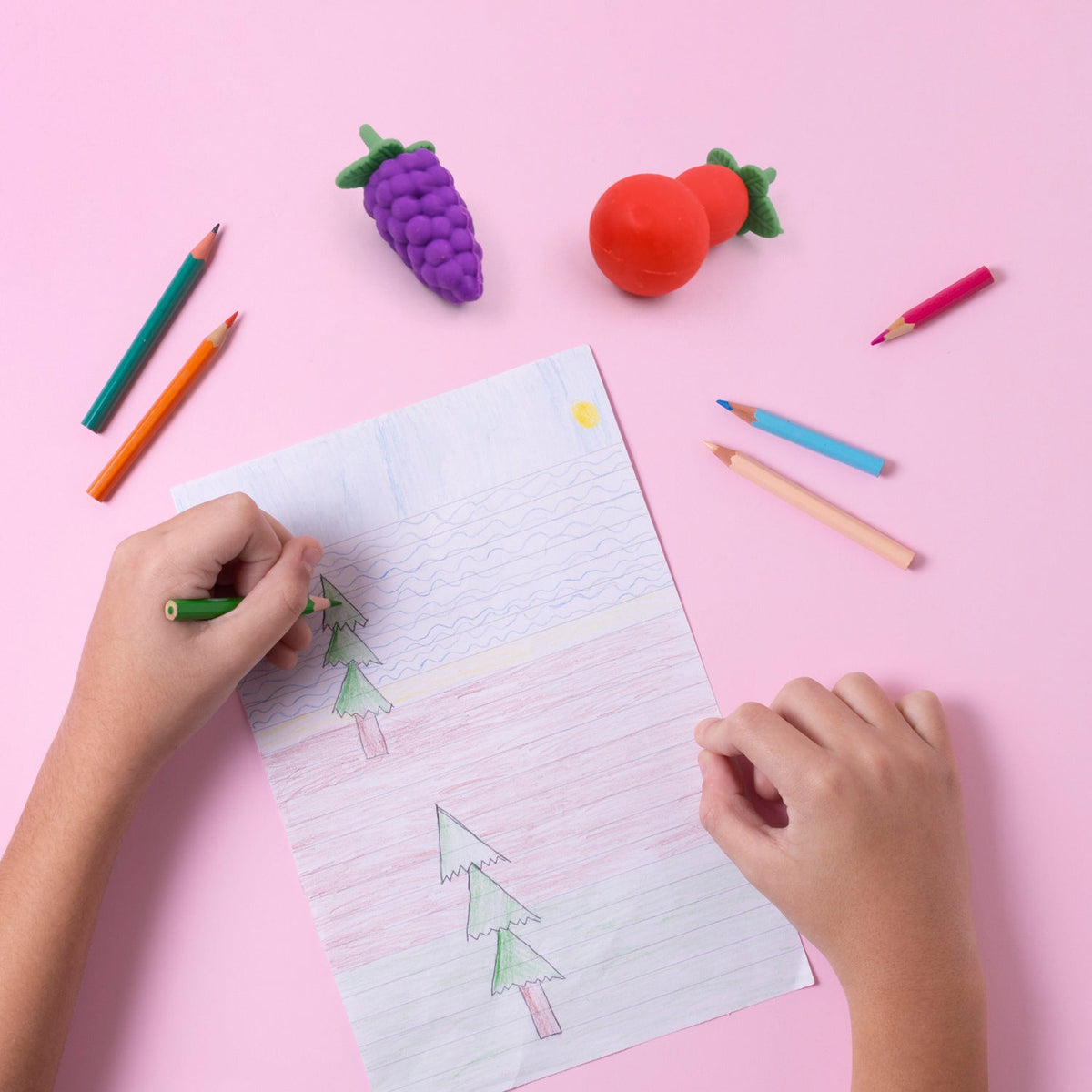 A child's hands color a drawing on a pink surface, with grape and cherry erasers nearby.
