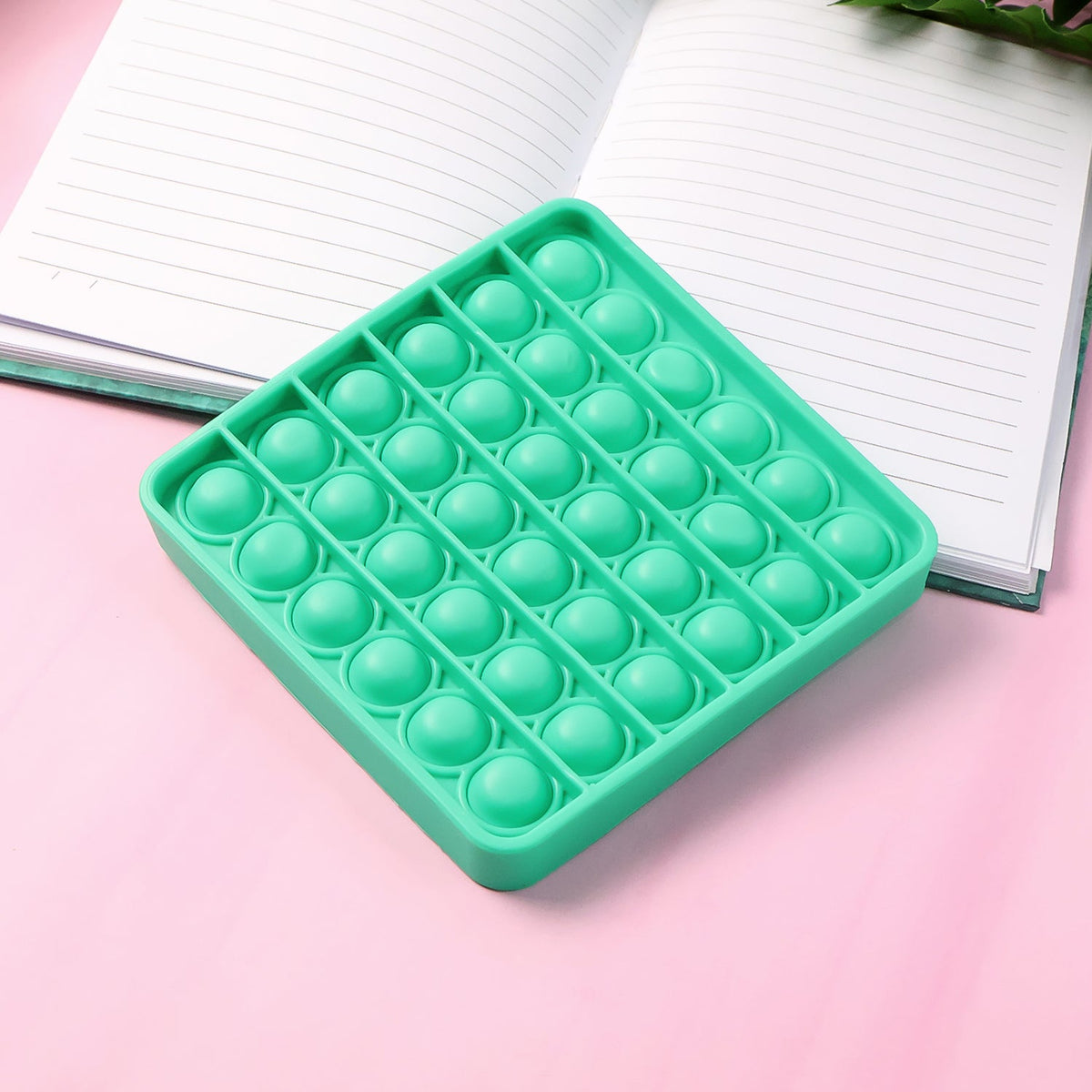 A square green silicone pop-it fidget toy resting on a pink surface next to an open notebook.