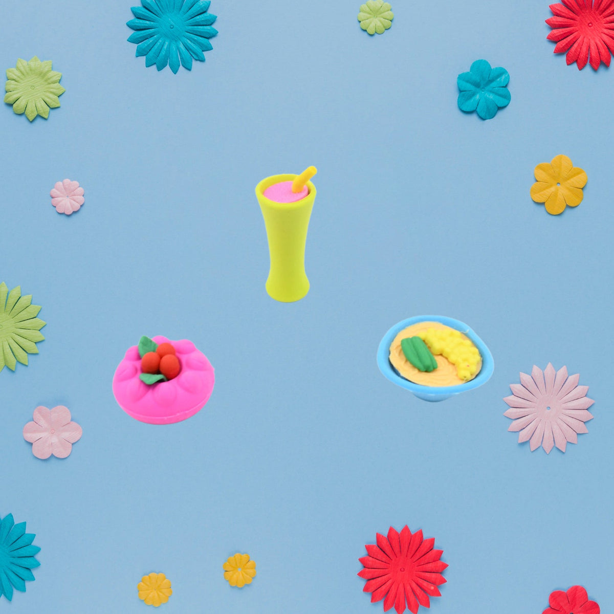 Three food-shaped erasers, a green drink, a pink cake, and a bowl of ramen, are centered on a blue background with colorful