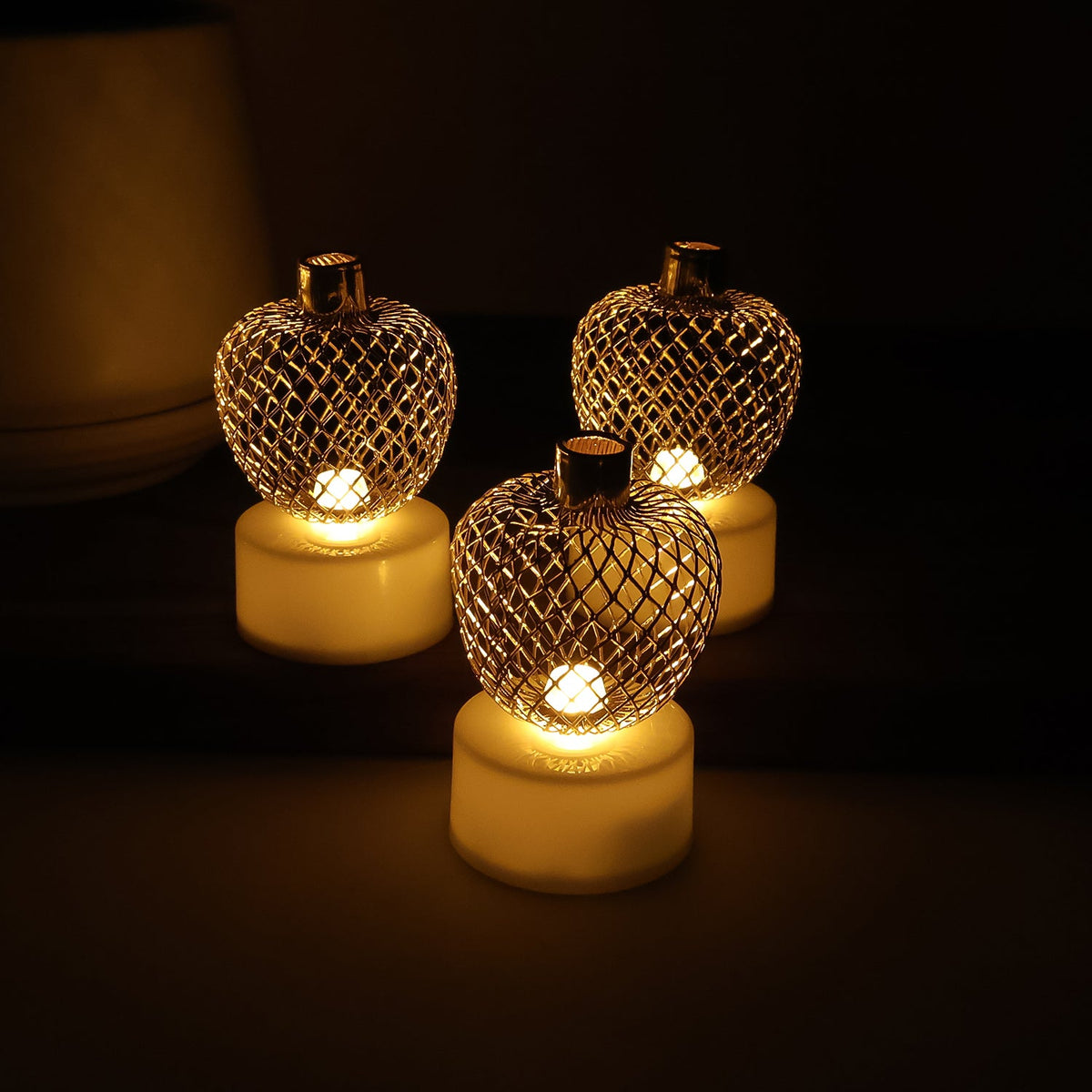 Three glowing gold mesh LED tealights arranged closely together on a dark surface, casting a warm glow.