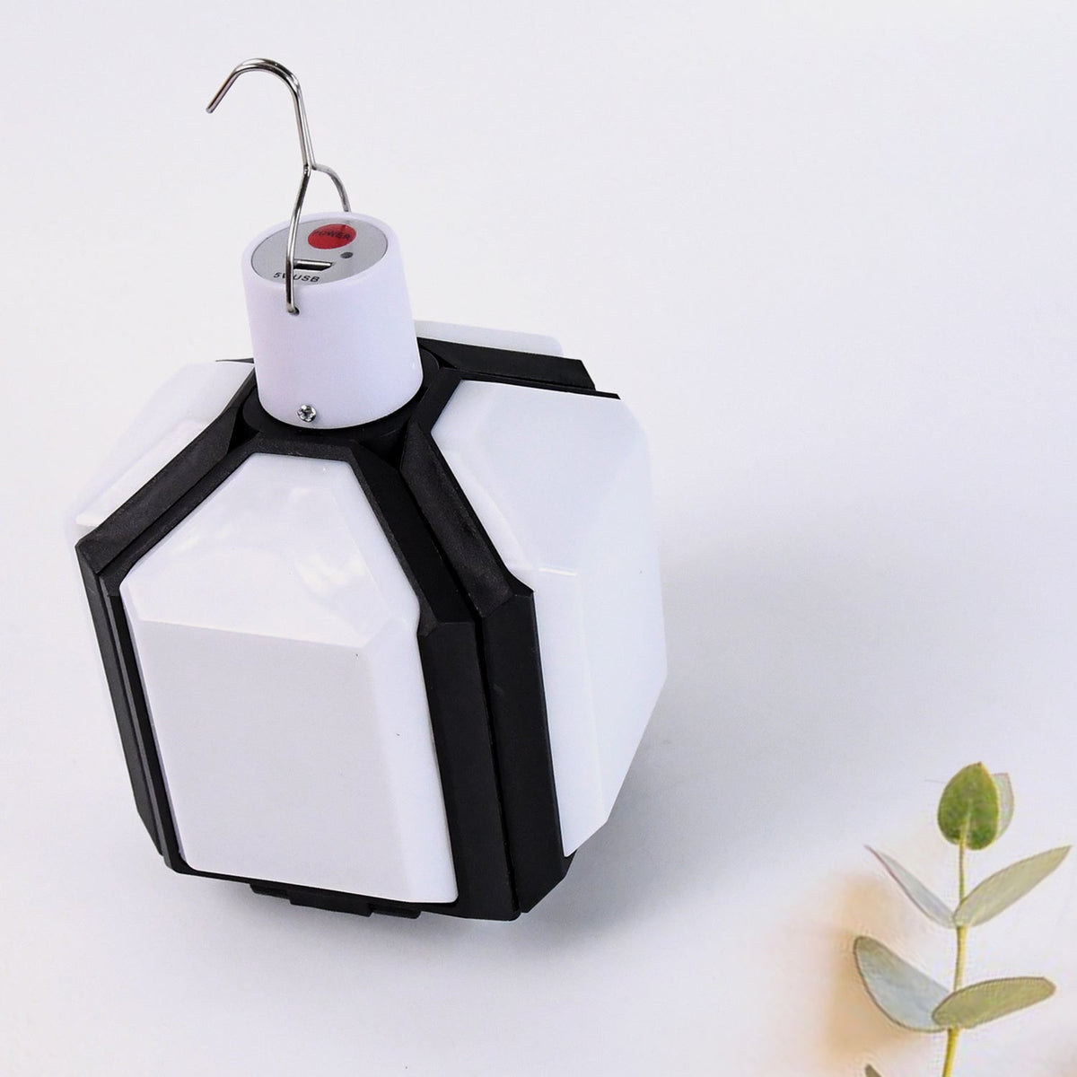 White and black foldable LED lantern in its compact form, with a plant sprig on a white background.
