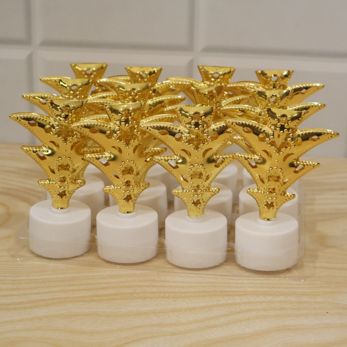A set of twelve golden Christmas tree LED tealight candles are packaged in clear plastic on a wooden surface.