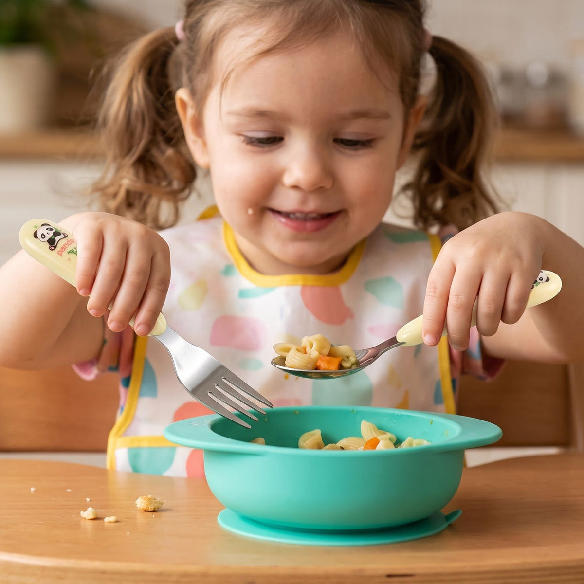 A young child with pigtails uses a yellow panda spoon and fork to eat pasta from a teal silicone bowl.