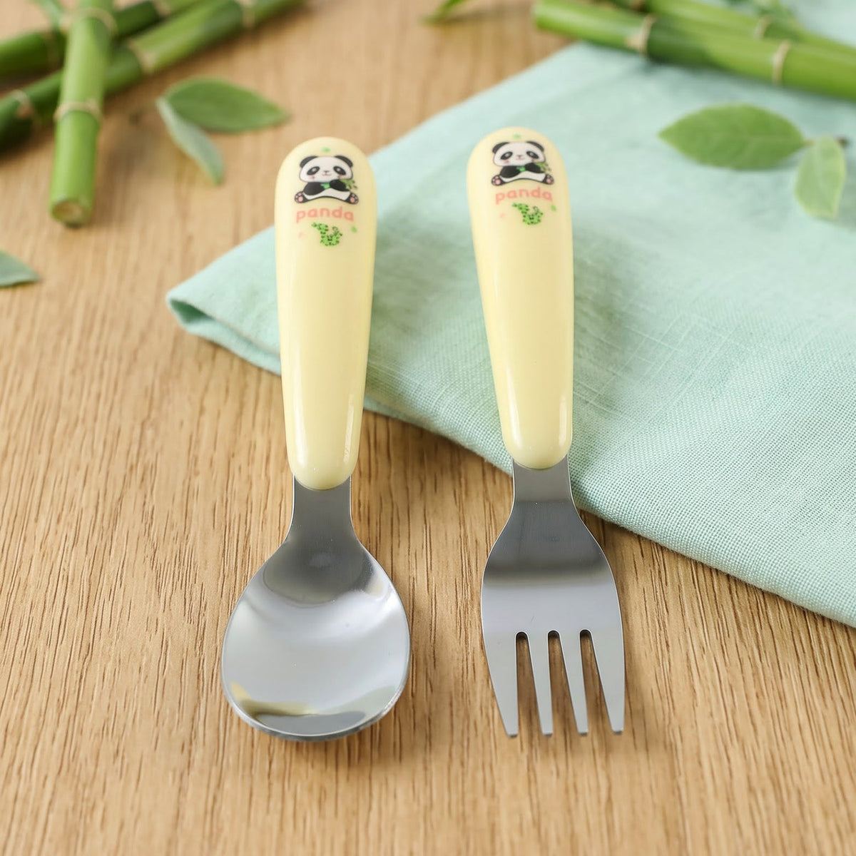 A yellow and silver panda-themed spoon and fork set rests on a light blue napkin with bamboo stalks in the background.