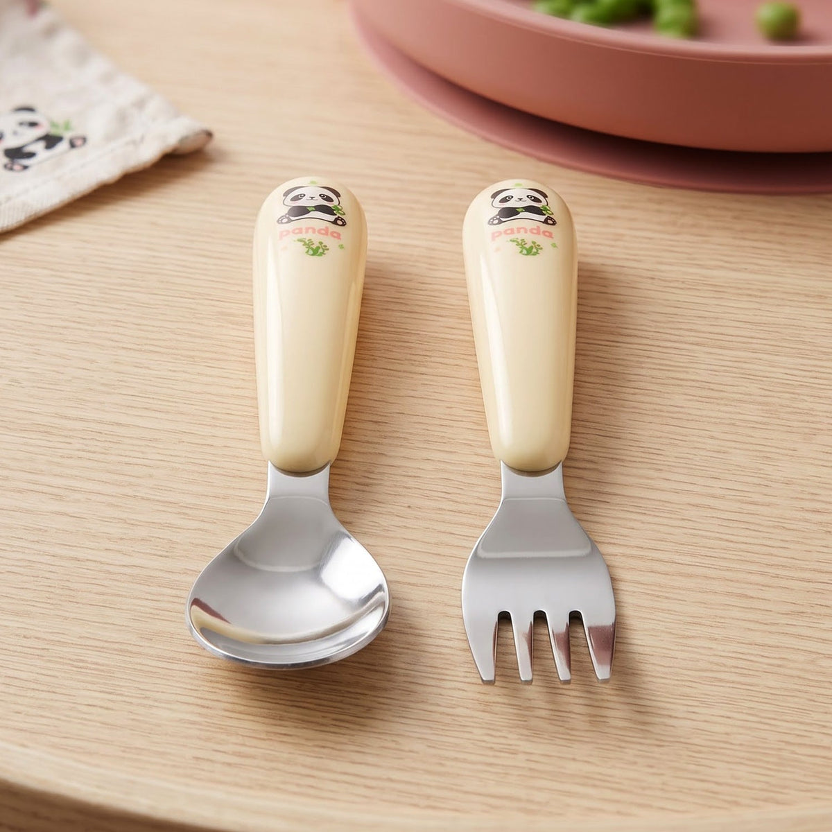 A yellow and silver panda-themed spoon and fork set rests on a wooden surface beside a pink plate.