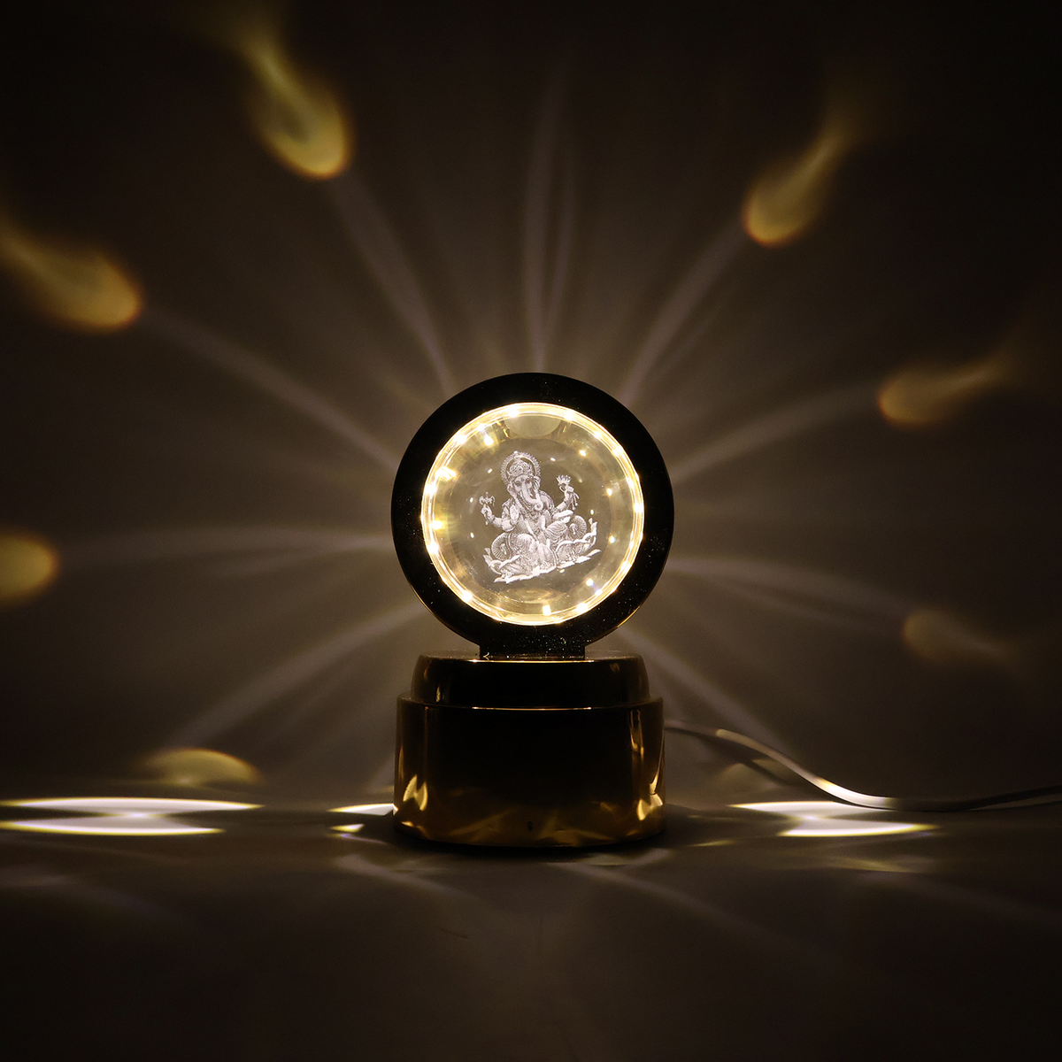 A golden lamp with a glowing Ganesha crystal projects starburst patterns onto a dark background, with a white cable.