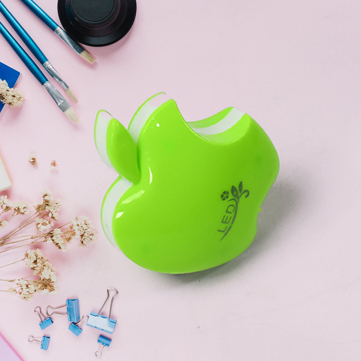 Top-down view of the green apple-shaped LED night light on a pink surface with art supplies and dried flowers.
