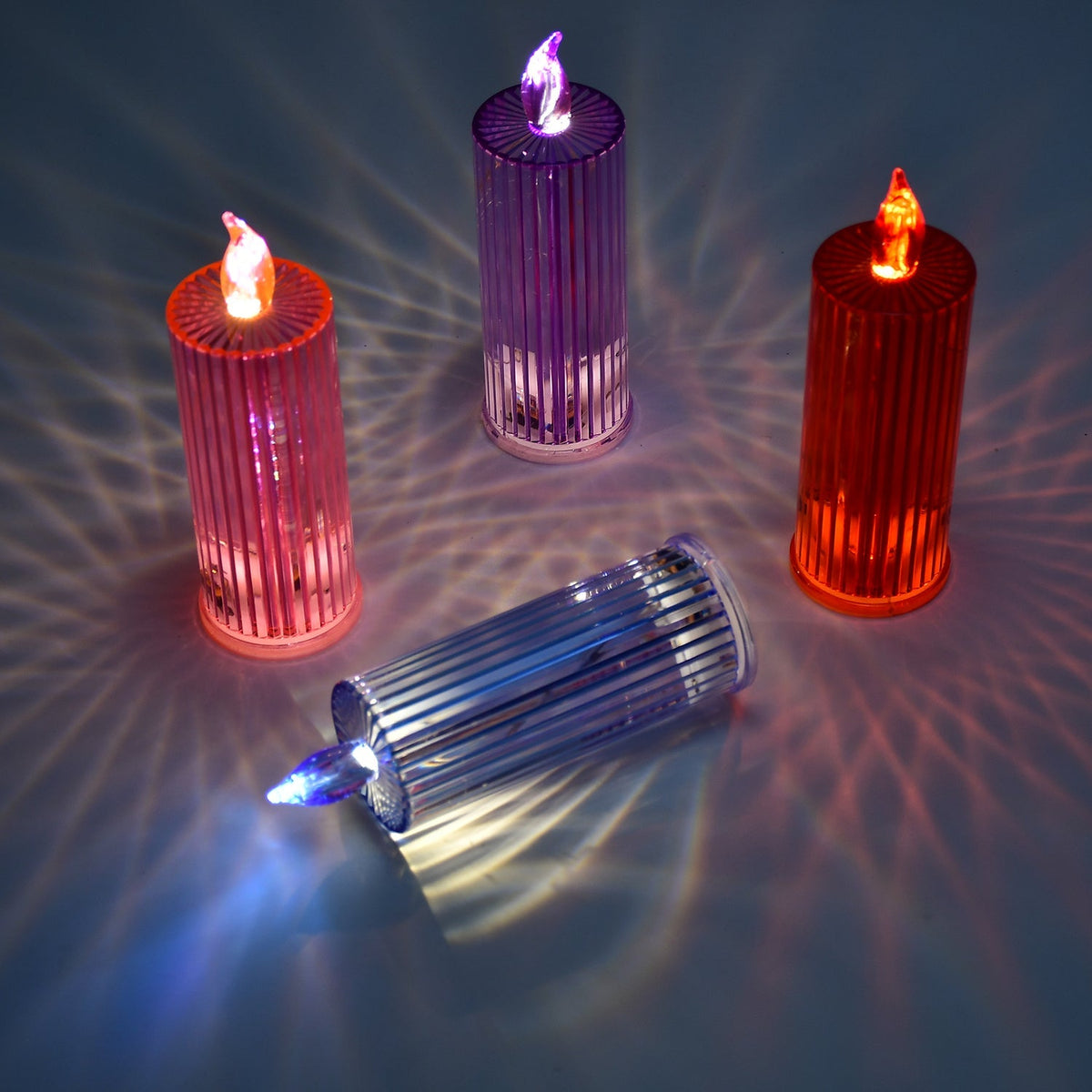 Three upright LED crystal candles in pink, purple, and orange, with one blue candle lying down, casting light patterns.