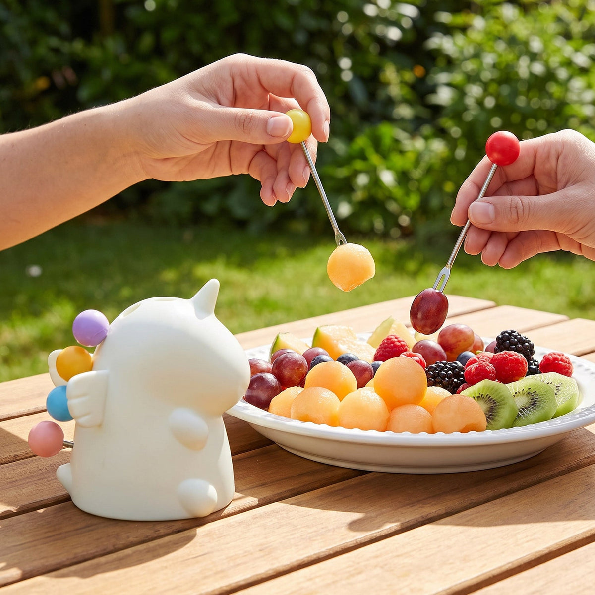 Two hands use colorful fruit forks to pick up melon and grapes from a white plate of mixed fruit outdoors.