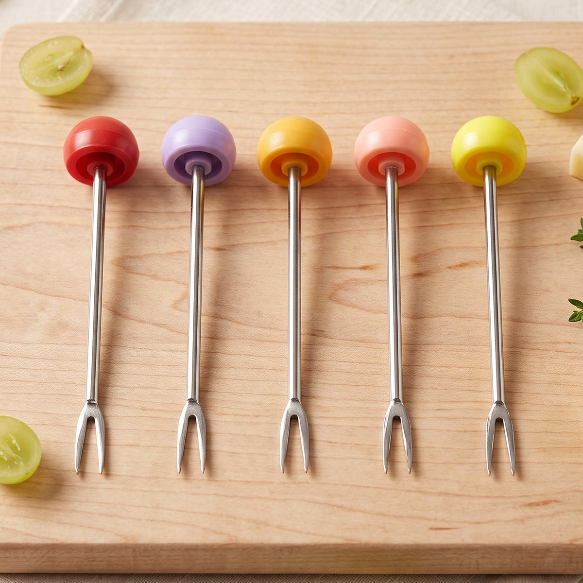Five stainless steel fruit forks with colorful spherical handles are arranged neatly on a light wooden cutting board.