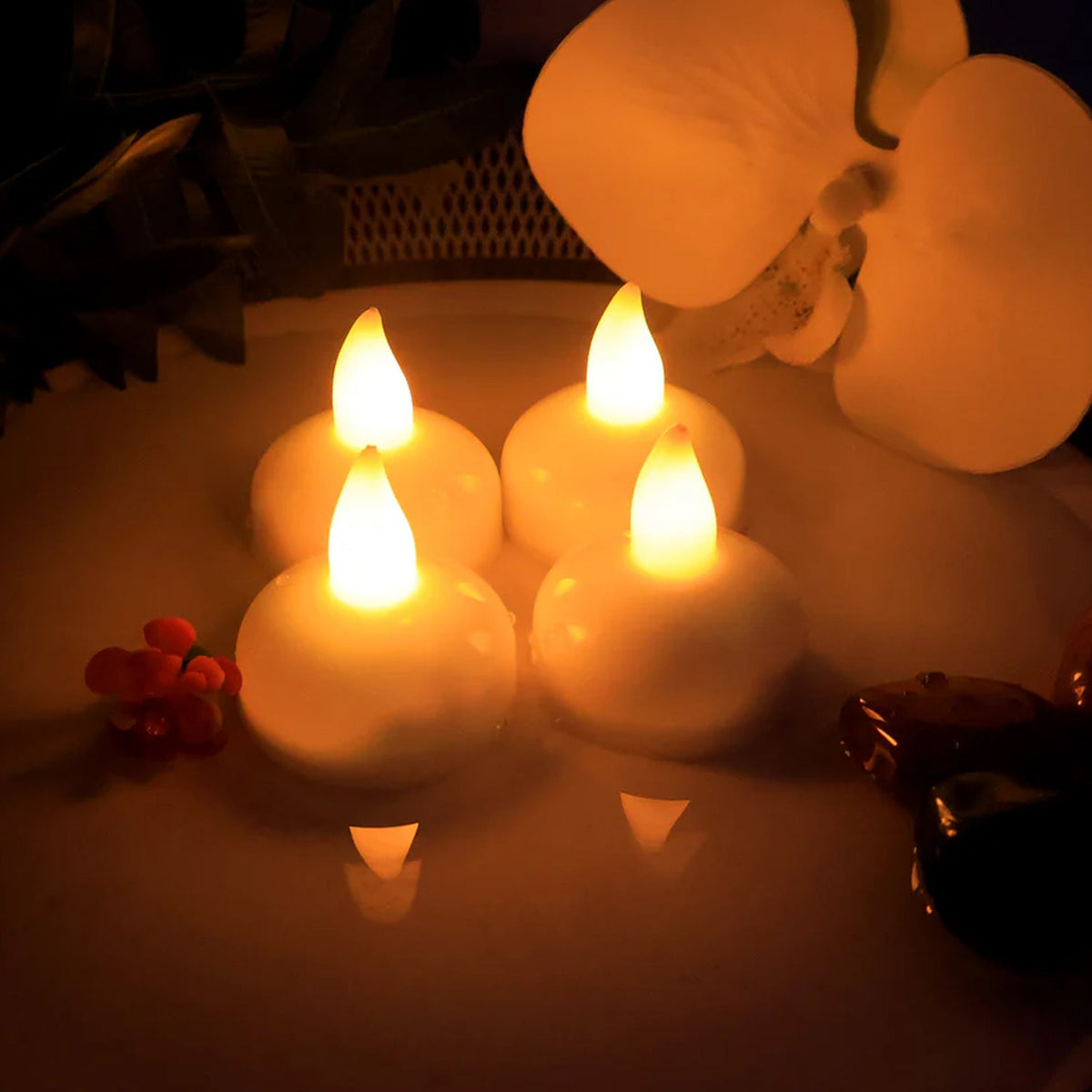 Four white LED tealight candles with warm yellow flames, floating in water, with white flowers in the background.