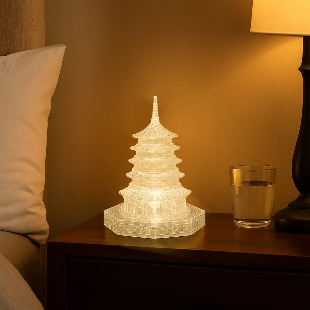 An illuminated LED crystal pagoda light on a wooden bedside table next to a glass of water and a lamp, with a white pillow.