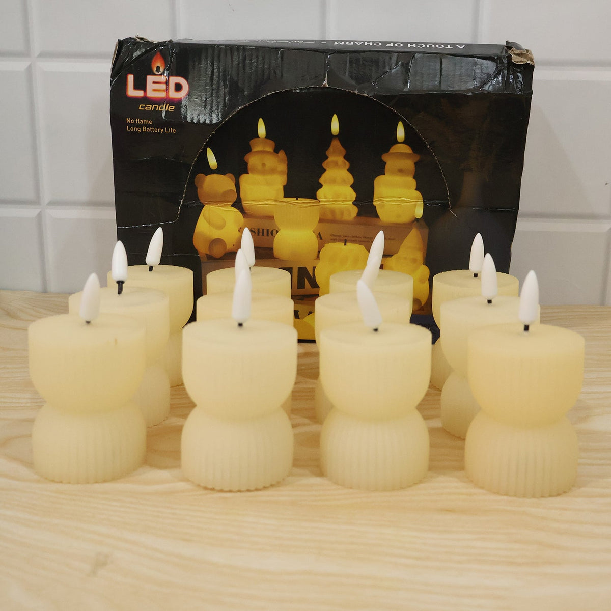 Twelve cream ribbed flameless candles with white wicks are arranged in front of their product box on a wooden surface.