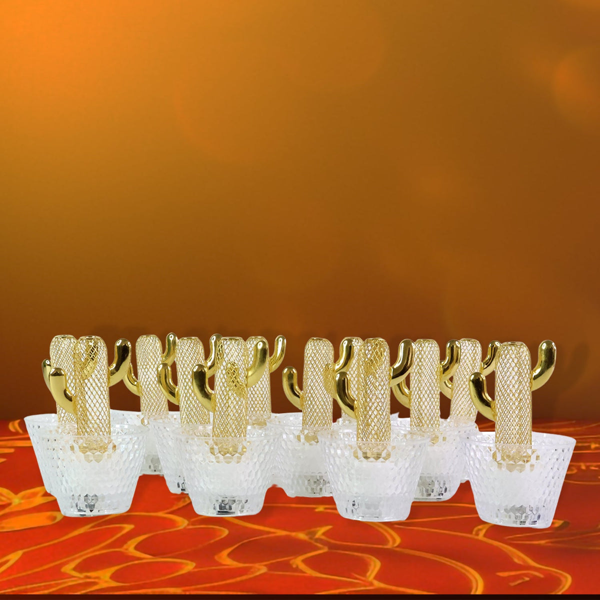 Twelve gold cactus LED tealights with clear bases glow on a red and orange patterned surface against an orange wall.