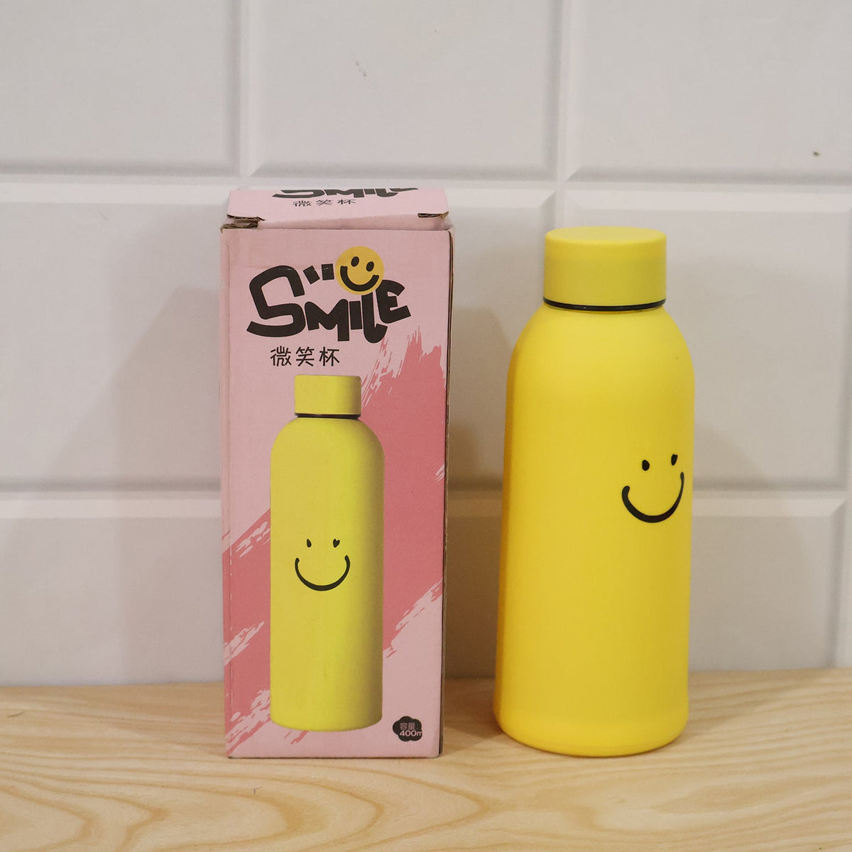 Yellow glass water bottle with a smiley face next to its pink packaging box, both featuring the same design.