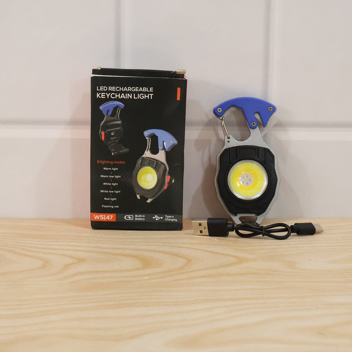 The keychain light, its USB-C cable, and its product box are arranged on a light wooden surface against a white wall.