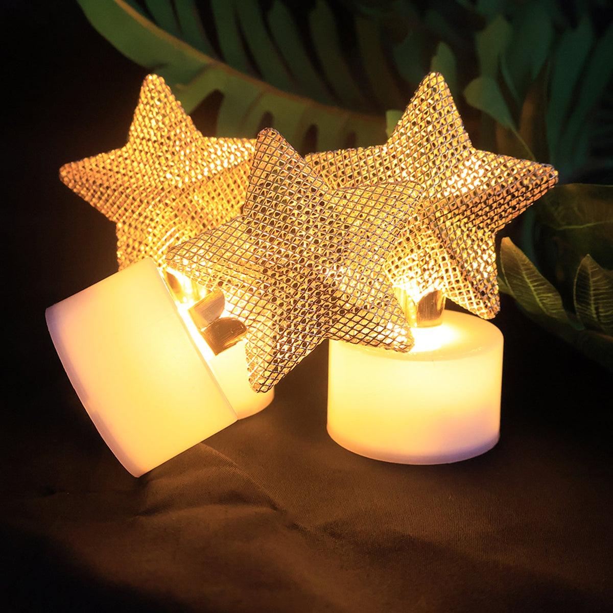 Three LED tealights with golden mesh stars, two standing and one tilted, glowing against dark foliage.